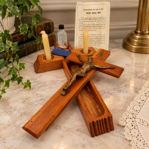 May include: A wooden sick call set featuring a crucifix with a bronze-toned figure, two cream-colored candles in holders, and a small glass bottle. A paper instruction card titled Instructions for use of the SICK CALL SET is included.