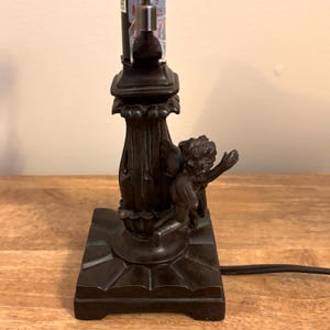 K&ouml;nnte beinhalten: Ein dunkelbrauner Lampenfu&szlig; aus Metall mit quadratischem Sockel und einer zentralen kannelierten S&auml;ule. Eine skulptierte Cherub-Figur mit Fl&uuml;geln ist an der Seite der S&auml;ule angebracht. Der Fu&szlig; umfasst ein schwarzes Stromkabel und eine Lampenfassung.