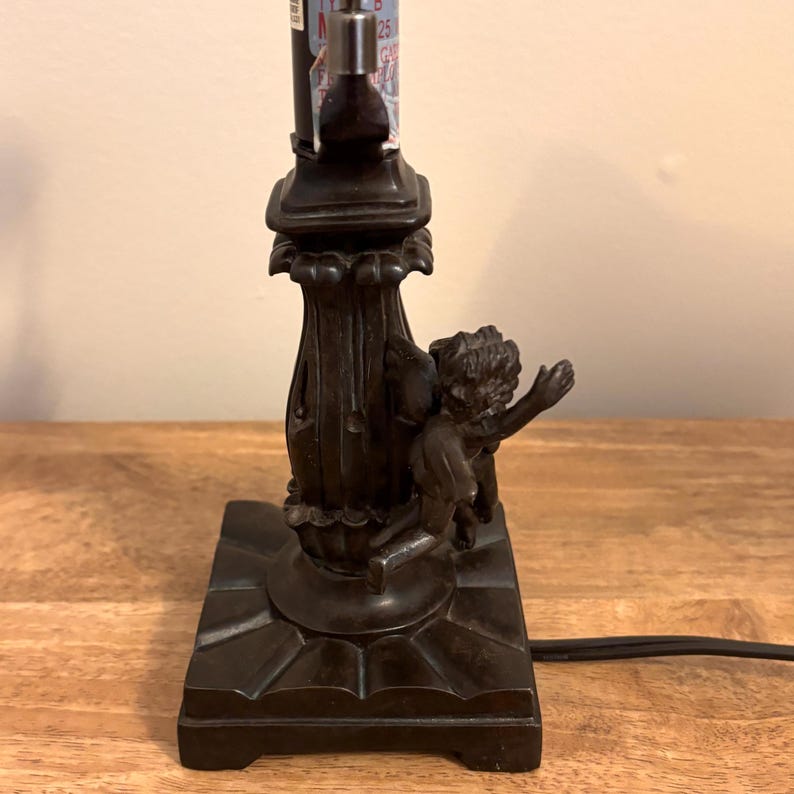 K&ouml;nnte beinhalten: Ein dunkelbrauner Lampenfu&szlig; aus Metall mit quadratischem Sockel und einer zentralen kannelierten S&auml;ule. Eine skulptierte Cherub-Figur mit Fl&uuml;geln ist an der Seite der S&auml;ule angebracht. Der Fu&szlig; umfasst ein schwarzes Stromkabel und eine Lampenfassung.