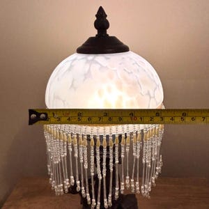 K&ouml;nnte beinhalten: Eine Tischlampe mit einem wei&szlig;en, marmorierten Glas-Kuppelschirm und einem dunkelbraunen Endst&uuml;ck. Ein gelbes Ma&szlig;band &uuml;ber dem Schirm zeigt einen Durchmesser von ca. 15 cm. Der Schirm hat einen Behang aus klaren und cremefarbenen Perlen.