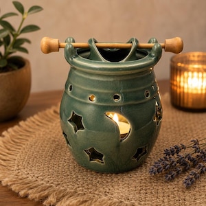 May include: A teal ceramic wax warmer featuring a glossy crackle glaze finish, cut-out star and crescent moon shapes, and a horizontal wooden rod resting across the top rim. The vessel has a rounded body with small circular ventilation holes.