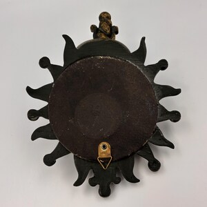 May include: A circular wall-mounted object with a black sunburst frame featuring pointed rays. The center is a dark brown circular panel. A gold-colored metal triangular hanging bracket is attached to the bottom, and a gold finial sits at the top.