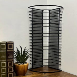 May include: Black metal corner shelf with a mesh design. The shelf has a curved top and a wooden base. A small potted plant and two books are visible on the wooden surface.