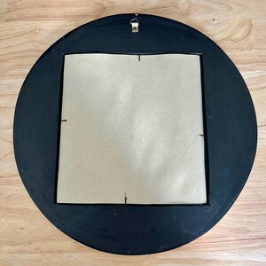 May include: Back of a round, black-painted wooden frame with a square cardboard backing. A metal hanger is attached at the top. The frame is on a light brown wooden surface.
