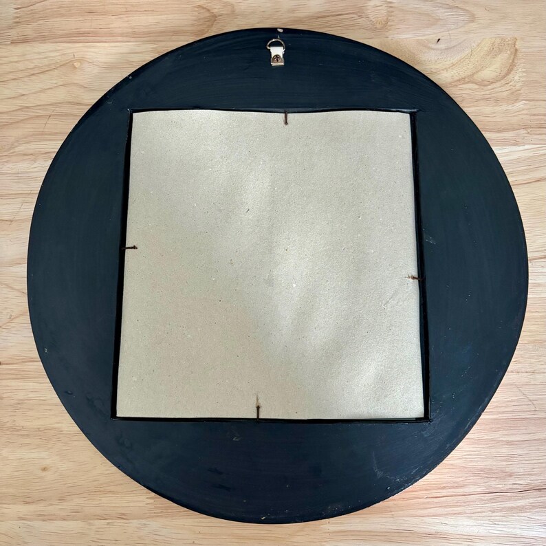 May include: Back of a round, black-painted wooden frame with a square cardboard backing. A metal hanger is attached at the top. The frame is on a light brown wooden surface.