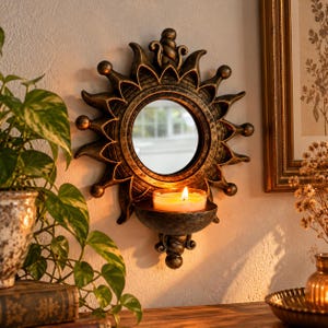 May include: A wall-mounted sunburst mirror featuring a dark bronze finish with gold highlights. The circular mirror is surrounded by radiating points and includes a small, integrated semi-circular shelf at the bottom holding a lit candle.