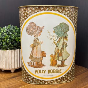 Peut inclure: Poubelle en métal vintage Holly Hobbie. La poubelle cylindrique présente un panneau central blanc avec une illustration circulaire de deux filles, un chien et un chat. La poubelle a un motif floral marron en haut et en bas, avec les mots "HOLLY HOBBIE" en or.