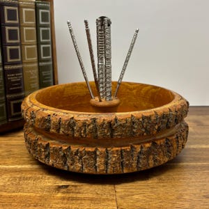 Puede incluir: Un cuenco de nueces de madera vintage con un exterior texturizado en forma de corteza. El cuenco contiene un cascanueces plateado y tres púas plateadas. El cuenco está hecho de madera marrón claro y se asienta sobre una superficie de madera. El fondo incluye una fila de libros.