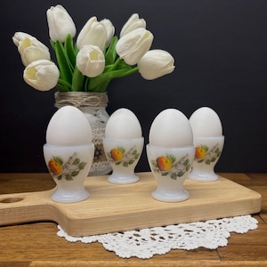 May include: Four white eggs in decorative egg cups with a painted fruit design, arranged on a wooden cutting board. A bouquet of white tulips in a rustic vase is in the background. The egg cups have a vintage aesthetic.
