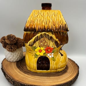 Peut inclure: Un pot à biscuits en céramique en forme de chaussure avec un toit de chaume. La chaussure est jaune avec une porte marron foncé et une petite fenêtre. Elle présente des fleurs peintes en jaune, orange et blanc sur le côté.