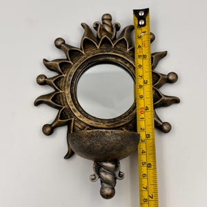 May include: A sun-shaped wall mirror with a bronze-toned finish and a small semi-circular shelf at the base. A yellow measuring tape is placed vertically against the mirror, showing a total height of approximately 7.5 inches.