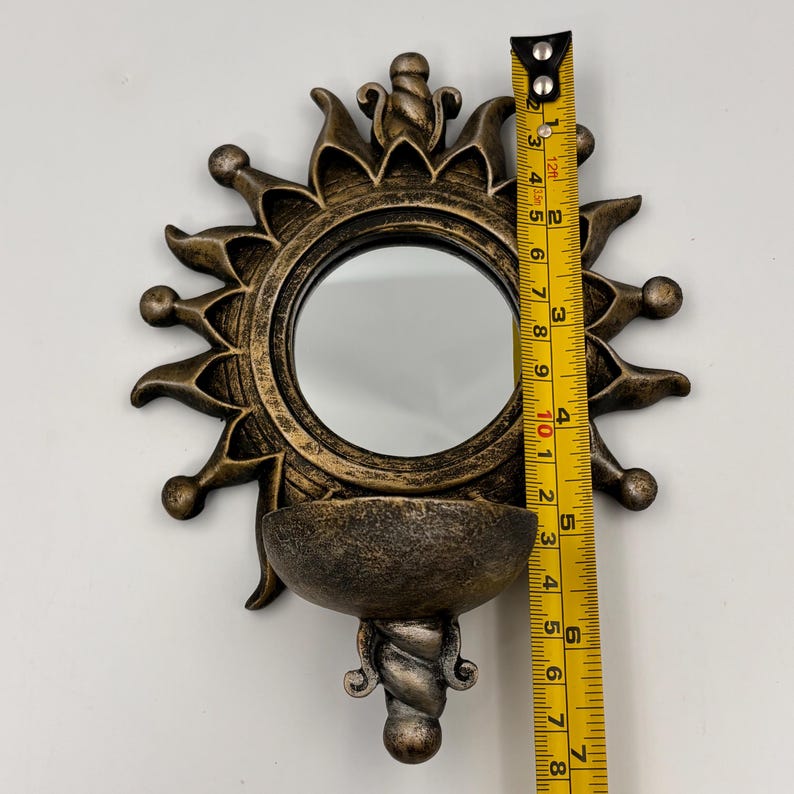 May include: A sun-shaped wall mirror with a bronze-toned finish and a small semi-circular shelf at the base. A yellow measuring tape is placed vertically against the mirror, showing a total height of approximately 7.5 inches.