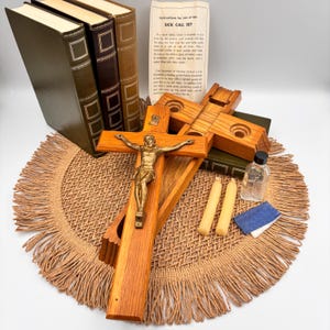 May include: A wooden sick call set featuring a crucifix with a brass-toned figure, two small candles, a glass holy water bottle, a blue cloth, and an instruction card. The set is displayed with three hardcover books in green, brown, and tan.