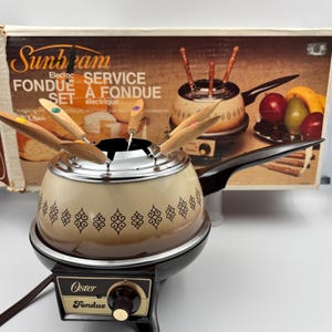 May include: An Oster electric fondue pot with a cream and brown gradient finish, featuring a black base with a temperature control knob and a metal rim. The pot includes a set of fondue forks with wooden handles and colored tips.