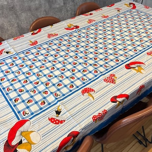 May include: A rectangular tablecloth featuring a central blue and white checkered pattern surrounded by a border of blue stripes. The fabric is decorated with a repeating print of red mushrooms with white spots and yellow stems throughout.