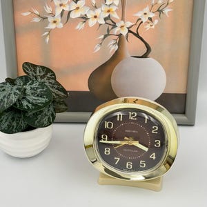 May include: A gold-toned Westclox Baby Ben alarm clock with a dark brown circular face, gold numerals, and gold hands. The clock features a polished metallic frame and a small base, with the text Baby Ben and Westclox printed on the dial.