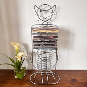 Op de afbeelding: Een zilverkleurig metalen cd-opbergrek in de vorm van een kat, met een draadframe kop inclusief snorharen en een spiraalvormig lichaam. Het rek bevat een stapel van acht cd-doosjes, elk met zichtbare tekst op de ruggen, rechtopstaand.