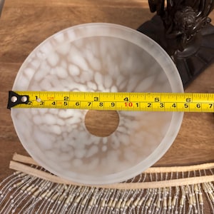 K&ouml;nnte beinhalten: Ein Lampenschirm aus Milchglas mit wei&szlig;em Fleckenmuster und einer zentralen runden &Ouml;ffnung. Ein gelbes Ma&szlig;band liegt oben auf und zeigt einen Durchmesser von etwa 17 cm. Darunter ist eine Perlenfransenborte zu sehen.