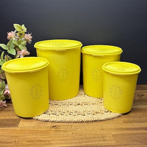 Può includere: Quattro contenitori di plastica gialli con coperchi. Ogni contenitore ha un motivo floreale decorativo e si trova su un tappetino intrecciato. I contenitori sono di diverse dimensioni.