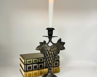 May include: A bronze-toned metal candlestick holder featuring a relief design of grape clusters and leaves, supporting a single lit white taper candle. The candlestick stands in front of a stack of three hardcover books with green and gold spines.