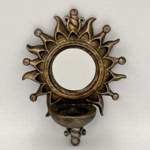 May include: A wall-mounted mirror featuring a circular glass center surrounded by a sunburst frame with a metallic bronze finish. Below the mirror is a small, semi-circular bowl-shaped shelf, also in a matching bronze tone.