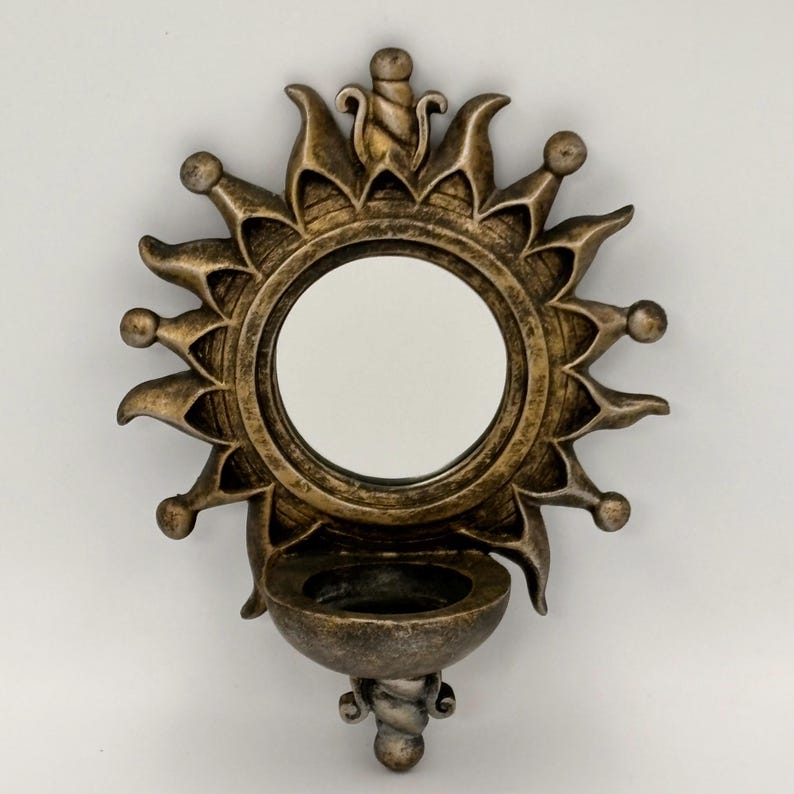 May include: A wall-mounted mirror featuring a circular glass center surrounded by a sunburst frame with a metallic bronze finish. Below the mirror is a small, semi-circular bowl-shaped shelf, also in a matching bronze tone.