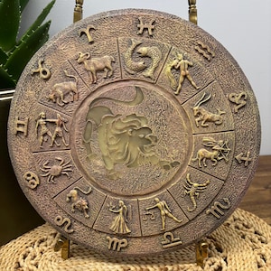 May include: A round, bronze-colored decorative plaque featuring zodiac symbols. The central image is a lion, surrounded by the twelve astrological signs. The plaque is displayed on a decorative stand, with a textured surface and gold accents.
