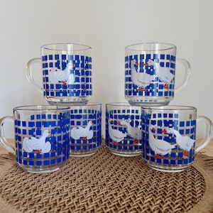 May include: Six clear glass mugs featuring a blue grid design and white goose illustrations. Each mug has a handle and a vintage aesthetic. The mugs are arranged on a woven surface, ideal for serving drinks. The mugs are a vintage design.