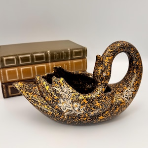 Könnte beinhalten: Ein schwanenförmiger Übertopf mit schwarzer Basis und goldenen Farbspritzern. Die Außenseite ist mit weißen Blumenillustrationen und kleinen klaren Kristallen verziert. Der Schwan hat einen gebogenen Hals und eine hohle Mitte.