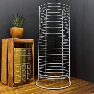 May include: A tall, white metal CD or DVD rack with a curved top and base. The rack is positioned next to a wooden crate holding books and a potted succulent. The background is a dark gray wall and a wooden floor.