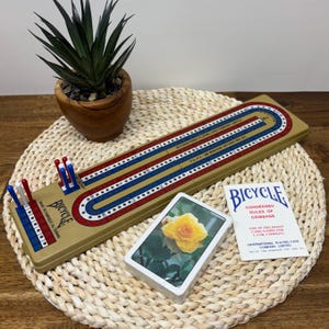 May include: A vintage cribbage board with red, white, and blue scoring tracks, a deck of Bicycle playing cards featuring a yellow rose, and a booklet titled "Condensed Rules of Cribbage". The board has a gold-coloured base.