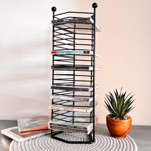 May include: A black metal CD rack filled with compact discs. The rack has a tall, vertical design with multiple shelves. A small potted plant sits to the right of the rack. CDs are visible on the bottom left.