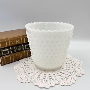 Könnte beinhalten: Eine weiße Hobnail-Vase mit einem gewellten Rand. Die Vase steht auf einer weißen gehäkelten Tischdecke. Im Hintergrund sind drei alte Bücher zu sehen.