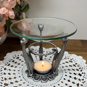 May include: A silver-coloured cat-themed candle holder with a clear glass dish. The candle holder features two cat figures supporting a circular ring, with a lit candle in the centre. The design includes floral accents and a decorative base.