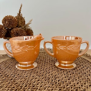 Peut inclure: Un ensemble de crème et sucrier en verre iridescent orange. Chaque pièce présente un motif de feuilles et deux anses. La crémière a un bec verseur. L'ensemble est présenté sur un tapis tissé.