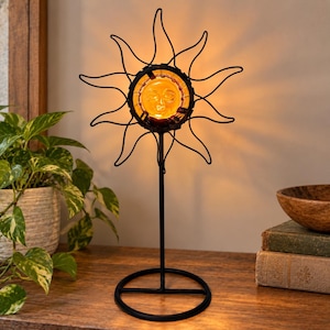 May include: A black metal sun-shaped lamp featuring a central amber glass disc with a molded face. The lamp has a thin vertical stand and a circular base, emitting a warm glow from the center, creating a sunburst silhouette with metal rays.