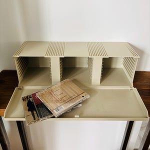 May include: A beige plastic CD storage rack with a hinged front door that folds down to reveal internal slots. The rack features vertical ribbed detailing on the top surface and holds a small stack of compact disc cases on the open door.