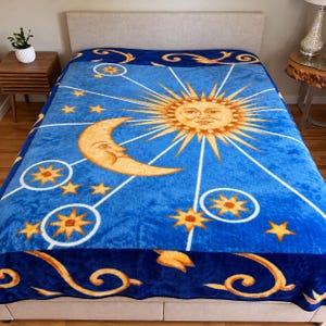May include: A plush blue blanket featuring a large golden sun with a face, a crescent moon, and various stars. The design includes white lines radiating from the sun and moon, with dark blue borders decorated with golden swirling patterns.
