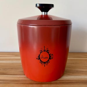 May include: A vintage red and orange grease container with a black handle and lid. The container has a decorative black emblem with the word "Grease" in the centre. The container is cylindrical with a slight taper and sits on a wooden surface.