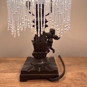 K&ouml;nnte beinhalten: Eine Tischlampe mit einem dunkelbraunen Sockel, der mit skulptierten Puttenfiguren verziert ist. Der Lampenschirm ist mit langen, herabh&auml;ngenden, klaren Glasperlen besetzt. Ein schwarzes Stromkabel f&uuml;hrt aus dem quadratischen Lampensockel heraus.
