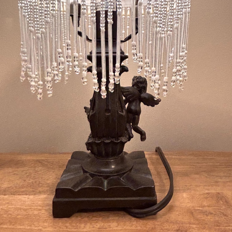 K&ouml;nnte beinhalten: Eine Tischlampe mit einem dunkelbraunen Sockel, der mit skulptierten Puttenfiguren verziert ist. Der Lampenschirm ist mit langen, herabh&auml;ngenden, klaren Glasperlen besetzt. Ein schwarzes Stromkabel f&uuml;hrt aus dem quadratischen Lampensockel heraus.