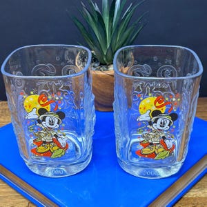 Puede incluir: Dos vasos cuadrados transparentes con un gráfico colorido de Mickey Mouse vestido con equipo de safari y binoculares. Los vasos están grabados con el año 2000 y patrones decorativos de estrellas y remolinos. Cada vaso tiene una base plana y esquinas redondeadas.