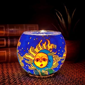 May include: A spherical glass candle holder featuring a vibrant blue background with a yellow sun and blue moon face design. The surface is decorated with yellow flames, green wave patterns, and small yellow stars scattered across the exterior.