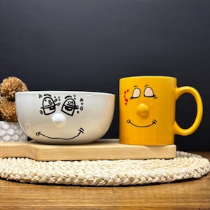 May include: A white ceramic bowl and a yellow mug with smiling faces. The bowl has black cartoon eyes and a smiling mouth. The yellow mug has a nose, eyes, and a smiling mouth. Both are on a wooden tray.