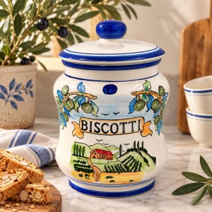 May include: A white ceramic canister with a blue lid, featuring hand-painted illustrations of olive branches and a landscape with a house. The word BISCOTTI is printed in black letters on a yellow banner across the centre of the jar.