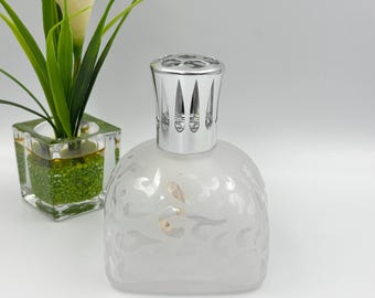 Könnte beinhalten: Eine Duftlampe aus mattiertem Glas mit einem abgerundeten, quadratischen Körper, der ein geätztes Wirbelmuster und eine polierte, silberfarbene Metallkappe aufweist. Die Lampe steht neben einer quadratischen Glasvase mit einer Grünpflanze.