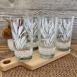 May include: Set of four clear drinking glasses with a white wheat stalk design. The glasses are cylindrical and sit on a wooden serving tray. The wheat design is repeated on each glass, with a decorative border at the base.