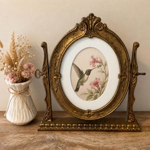 Peut inclure: Un cadre photo ovale pivotant doré avec des détails ornés, contenant une impression de colibri et de fleurs roses. À côté se trouve un vase en céramique blanche nervurée rempli d'un arrangement floral séché d'herbes beiges et roses.