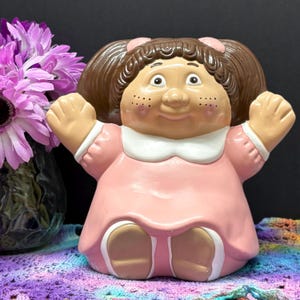 Peut inclure: Figurine de poupée en céramique vintage avec des cheveux bruns, des nœuds roses et une robe rose avec un col blanc. La poupée a des taches de rousseur et est posée avec les bras levés. Objet de collection.