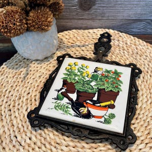 Op de afbeelding: Zwarte gietijzeren onderzetter met een keramische tegel met kruiden in potten en tuingereedschap. De tegel heeft een witte achtergrond met bruine potten, groene planten, gele bloemen en rode bessen. De onderzetter heeft een decoratief handvat.